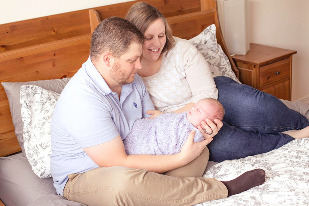father holds newborn baby wrapped up in the master bedroom with wife by his side